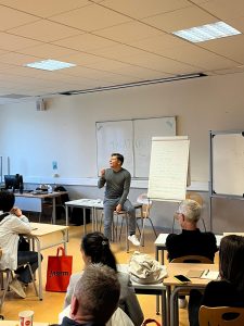 Salle de classe, avec un homme assis sur une table devant un tableau blanc.