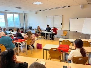 Salle de classe, une scénette de théâtre a lieu autour d'une table devant un tableau blanc.